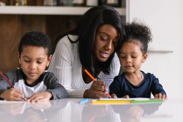 A mother helps her children with their lessons as she homeschool them during the pandemic. Both mixed race children of elementary school age are colouring in and seated at the kitchen island.