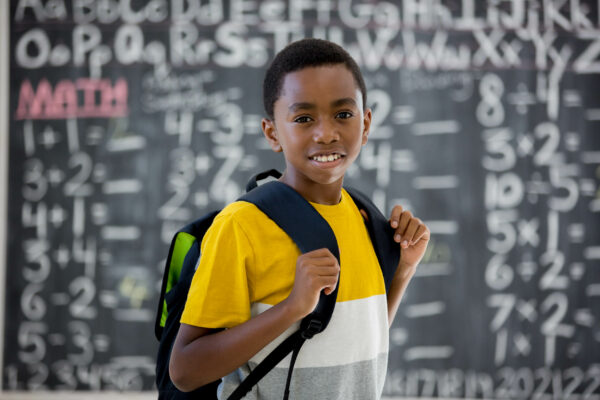 An elementary age schoolboy is standing at the front of the classroom on the first day of school and is smiling while looking at the camera.