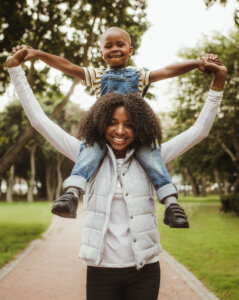 Happy mother giving son a piggyback ride on her shoulders in park. African woman carrying his boy on her shoulders while walking in a park.